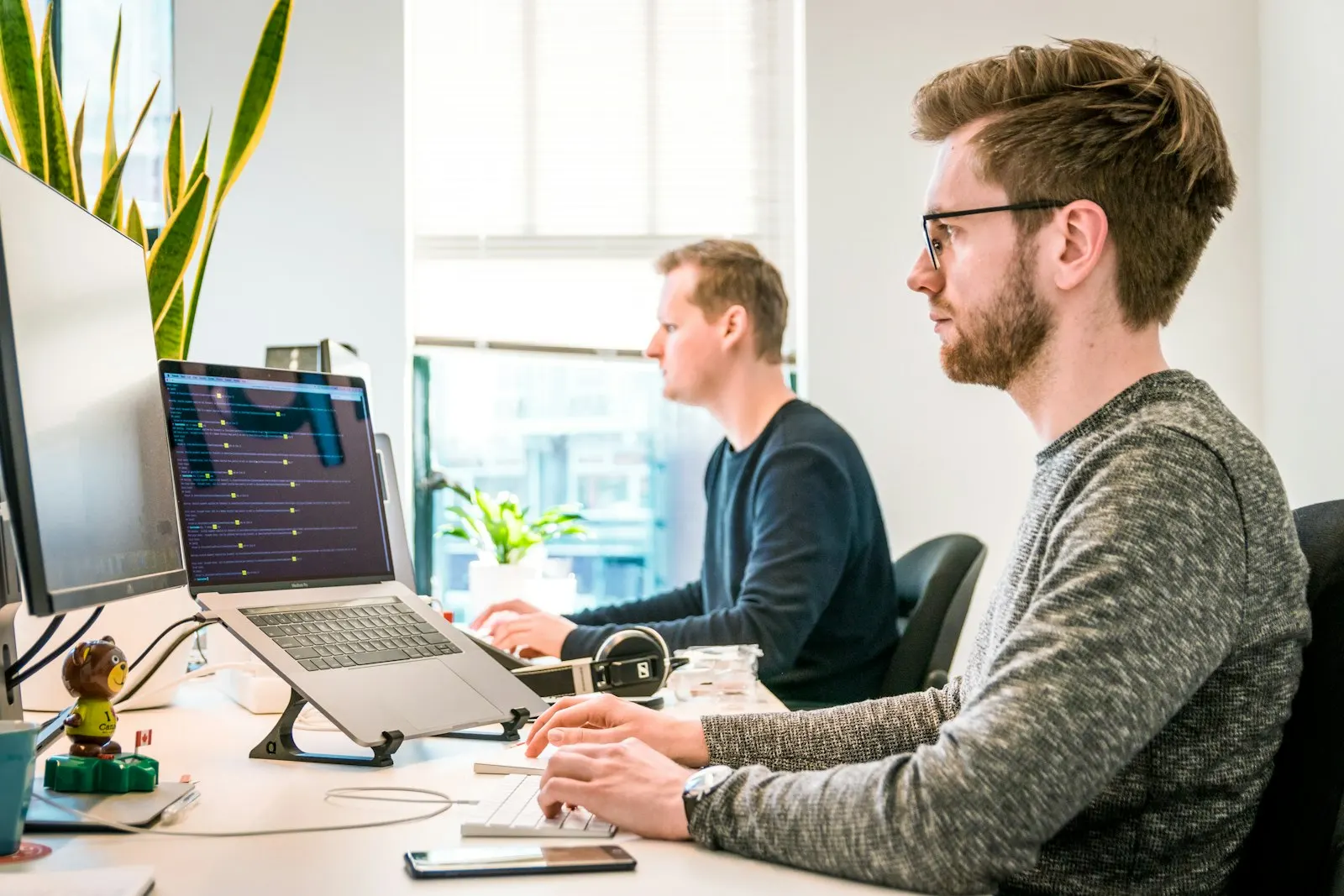An engineer writing code at a bright desk, with a teammate working in the background